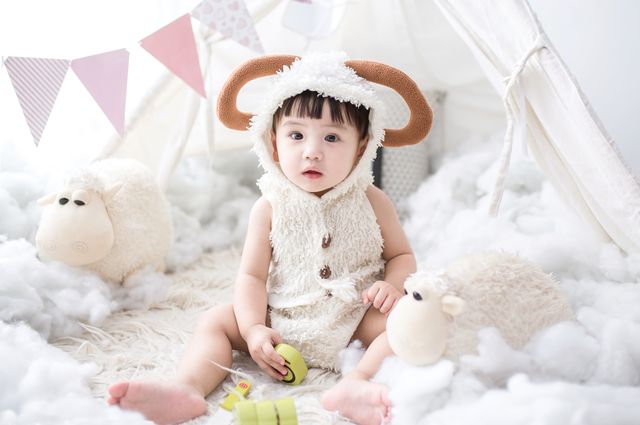 Best Baby Toys for Infants 0-6 Months, a baby sitting on a bed with white sheets, wearing a reindeer costume playing with some toys.